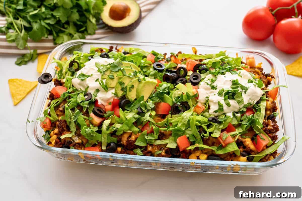 Taco Bake Recipe Served with Toppings A baked taco casserole, showcasing crispy tortilla chips, seasoned beef, beans, and corn, topped with melted cheese, and garnished with vibrant lettuce, ripe tomatoes, black olives, a swirl of sour cream, and fresh avocado slices.