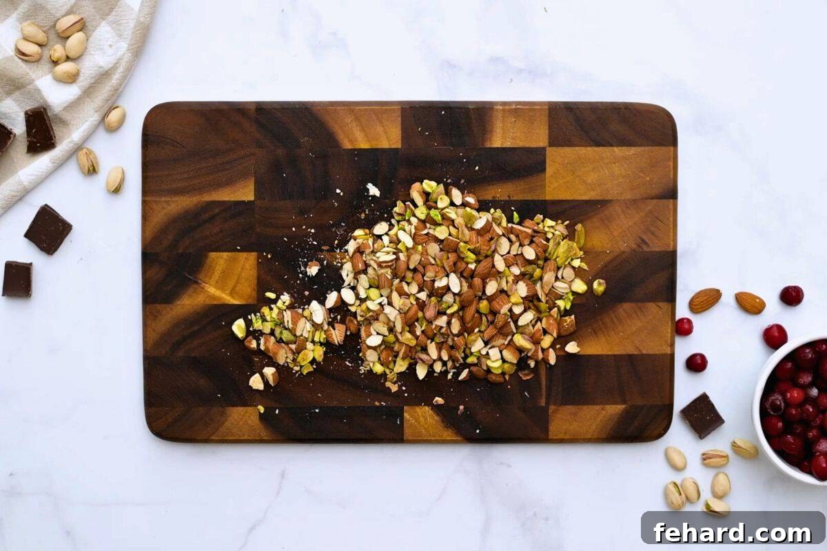 Freshly toasted and chopped pistachios and almonds are neatly arranged on a cutting board, alongside rich dark chocolate pieces, ready for the bark recipe.