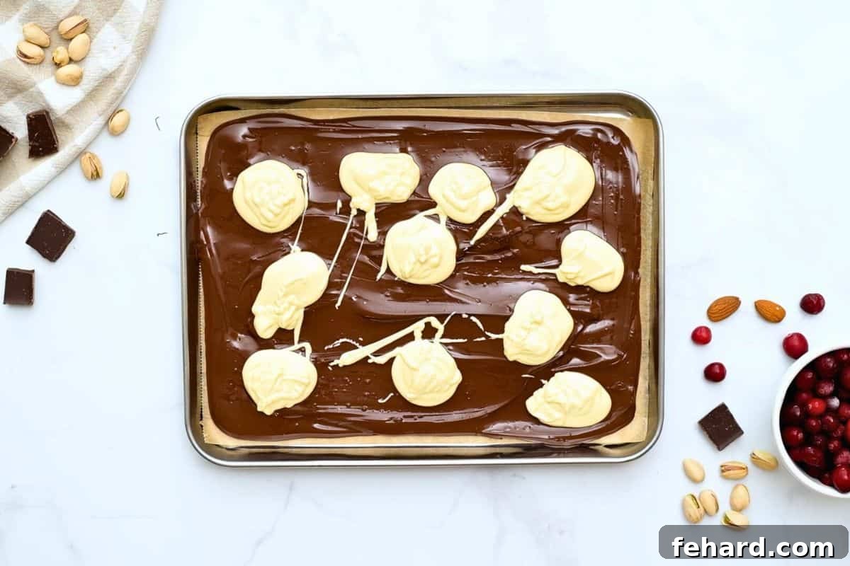 Delicate spoonfuls of melted white chocolate are artfully dolloped over a rich dark chocolate base on a parchment-lined baking sheet, preparing for the swirl.
