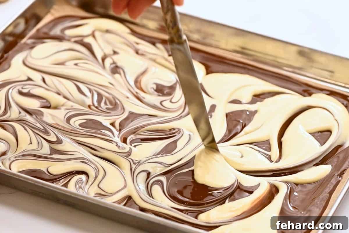 A butter knife gracefully swirls melted white chocolate into melted dark chocolate, crafting a beautiful marbled pattern for the bark.