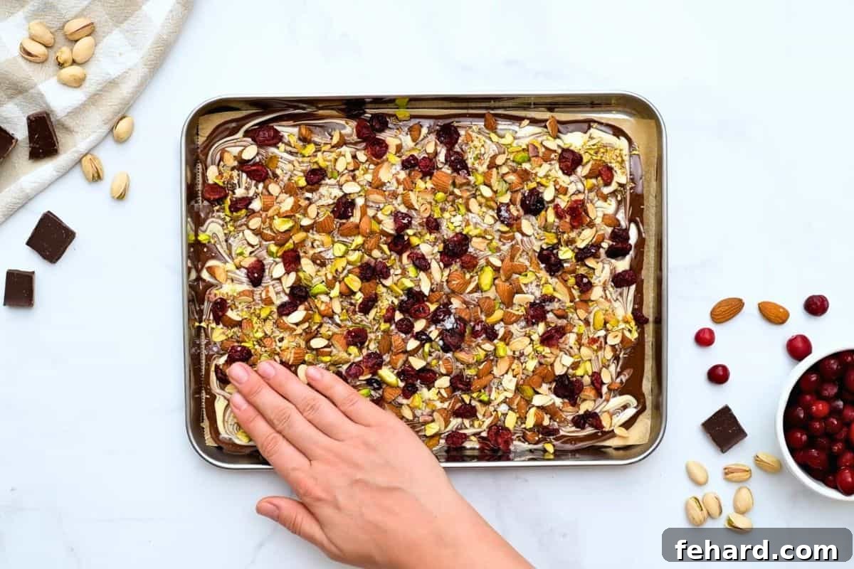 A hand delicately presses toasted nuts and dried cranberries into the soft, marbled chocolate bark, ensuring perfect adhesion.