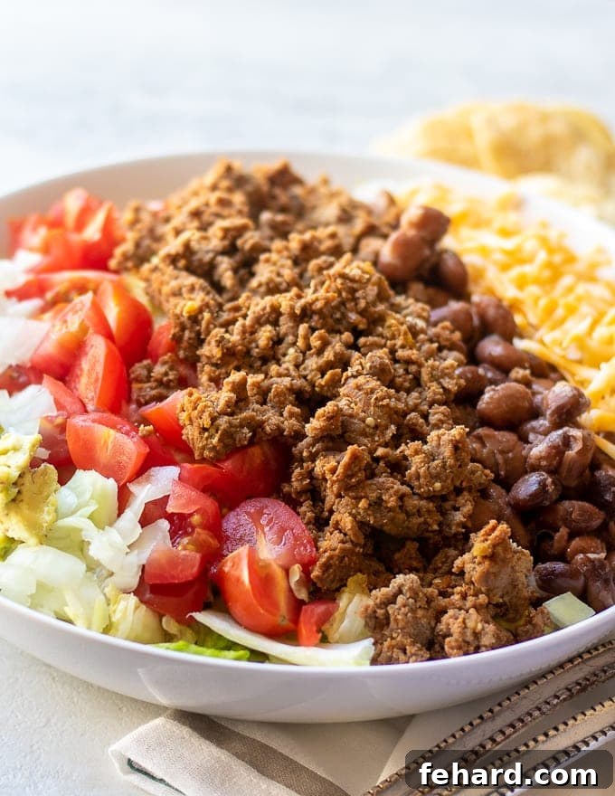 Delicious Homemade Taco Salad A crisp white bowl overflowing with layers of seasoned ground beef, vibrant diced tomatoes, shredded cheese, and tender beans, all nestled on a bed of fresh, crunchy lettuce – the perfect homemade Taco Salad.