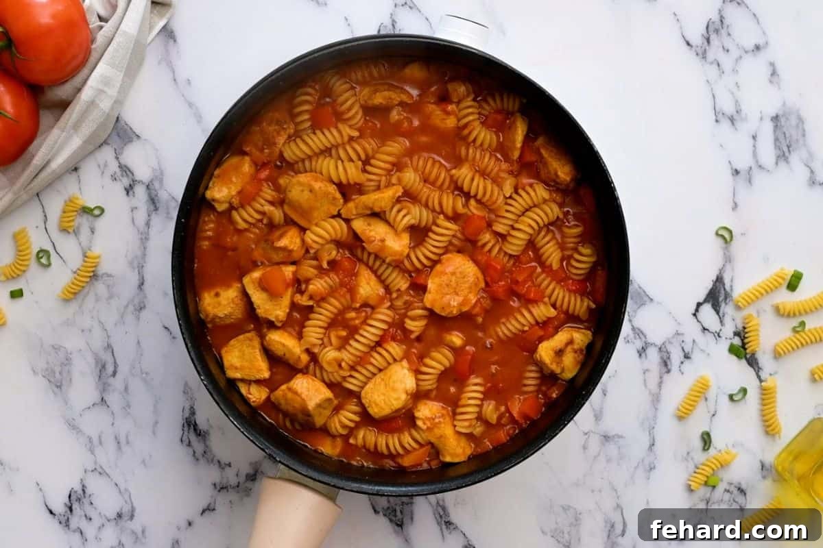 Uncooked rotini pasta, browned chicken, fire-roasted tomatoes, and chicken stock simmering together in a large skillet, absorbing rich Cajun flavors.