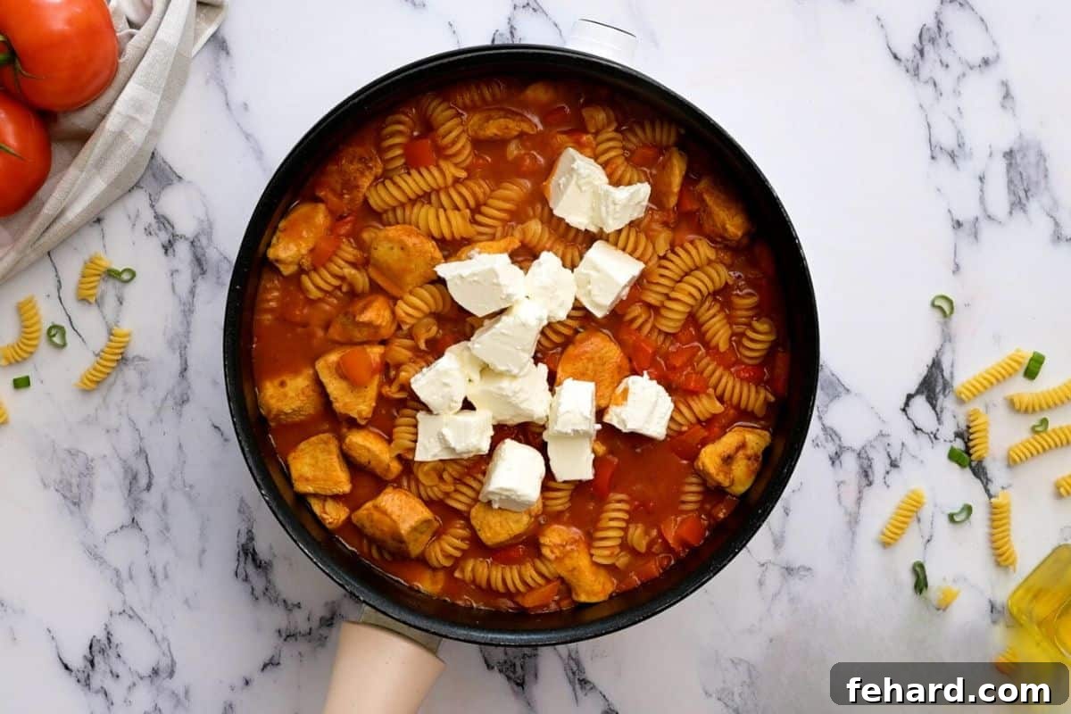 Cubed cream cheese being gently stirred into the hot, simmered Cajun pasta and chicken, beginning to melt and create a smooth, creamy sauce.