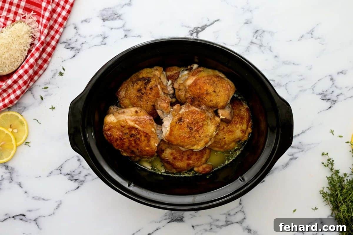 Seared chicken thighs arranged on top of a bed of seasoned rice, garlic, and chicken broth inside a slow cooker, ready for cooking.