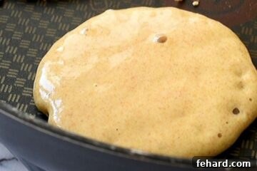 A whole wheat pancake cooking on a griddle, showing small, active bubbles on its surface.