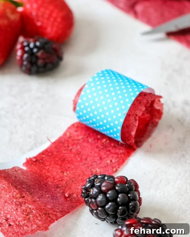 Homemade Fruit Roll-Ups Recipe A vibrant homemade fruit roll-up, elegantly rolled in blue craft paper, accompanied by fresh blackberries and strawberries, highlighting the natural goodness.