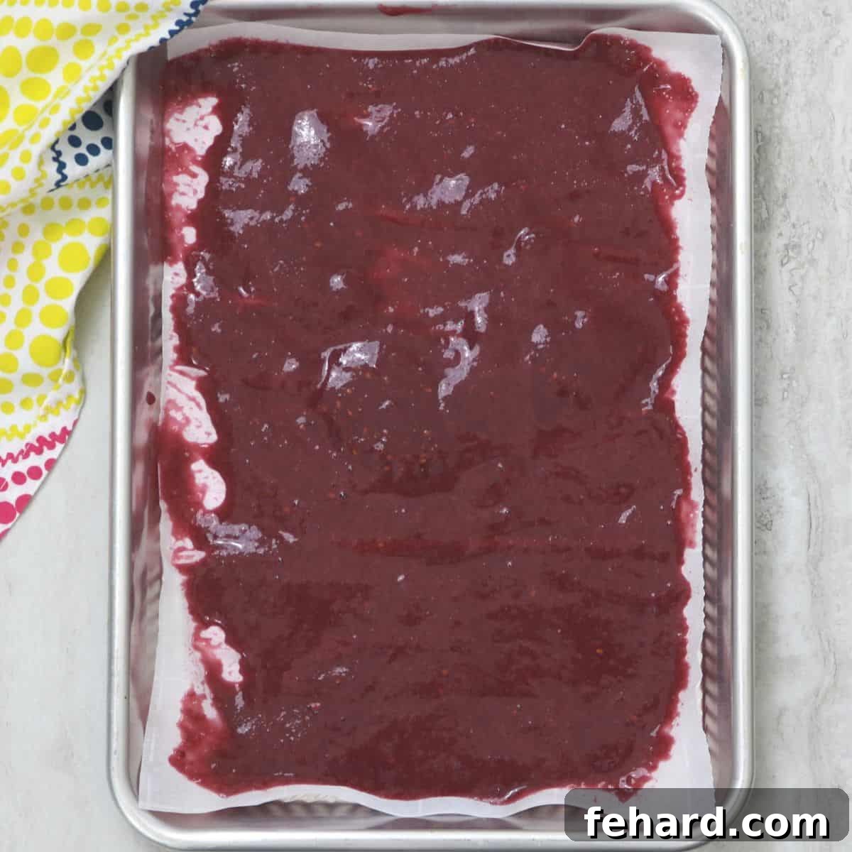 Copycat Fruit Roll-Ups Spreading A vibrant fruit roll-up mixture perfectly spread out on a sheet pan lined with parchment paper, ready for baking.