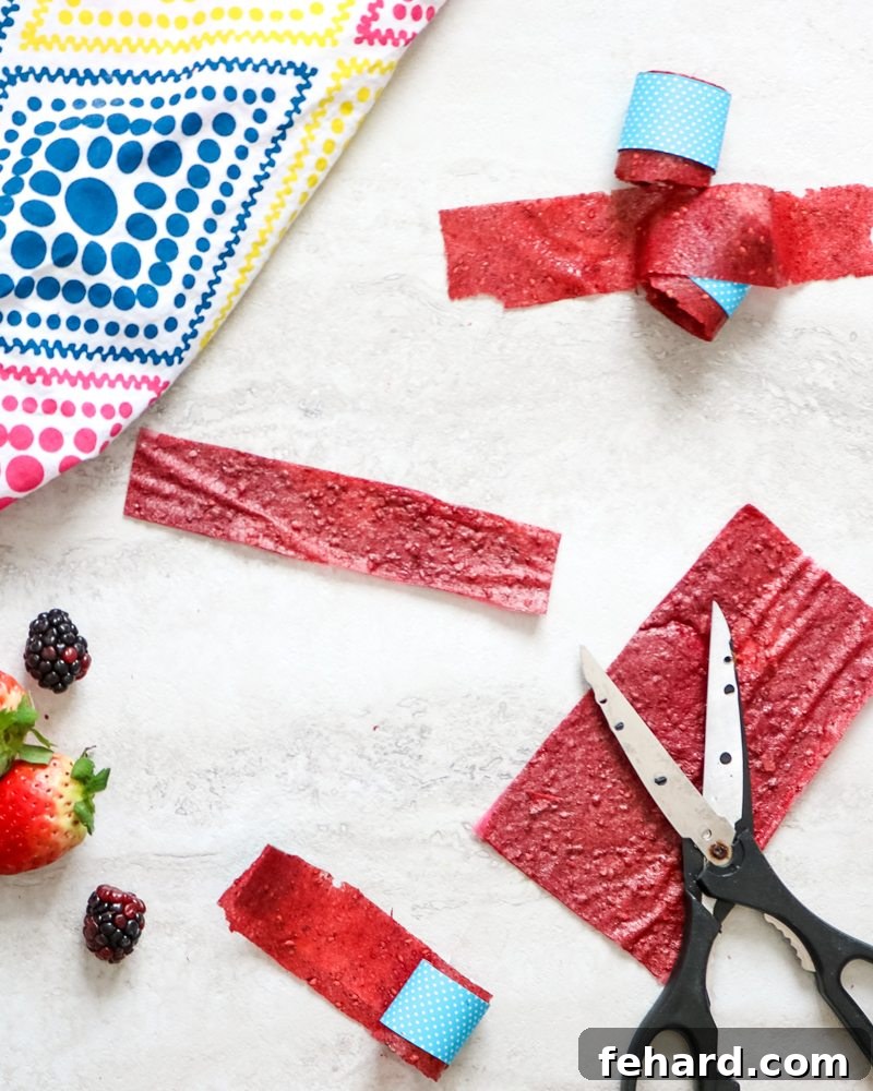 How to Make Fruit Roll-Ups Slicing Beautifully cut homemade strawberry fruit roll-ups arranged on a white board, surrounded by fresh fruit and kitchen scissors, ready to be rolled and enjoyed.