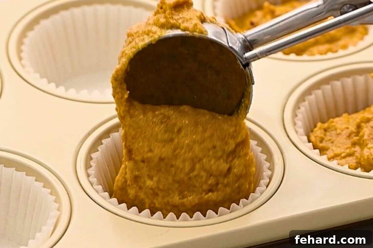 An ice cream scoop portioning pumpkin muffin batter into a lined muffin tin, ensuring even distribution for uniform baking.