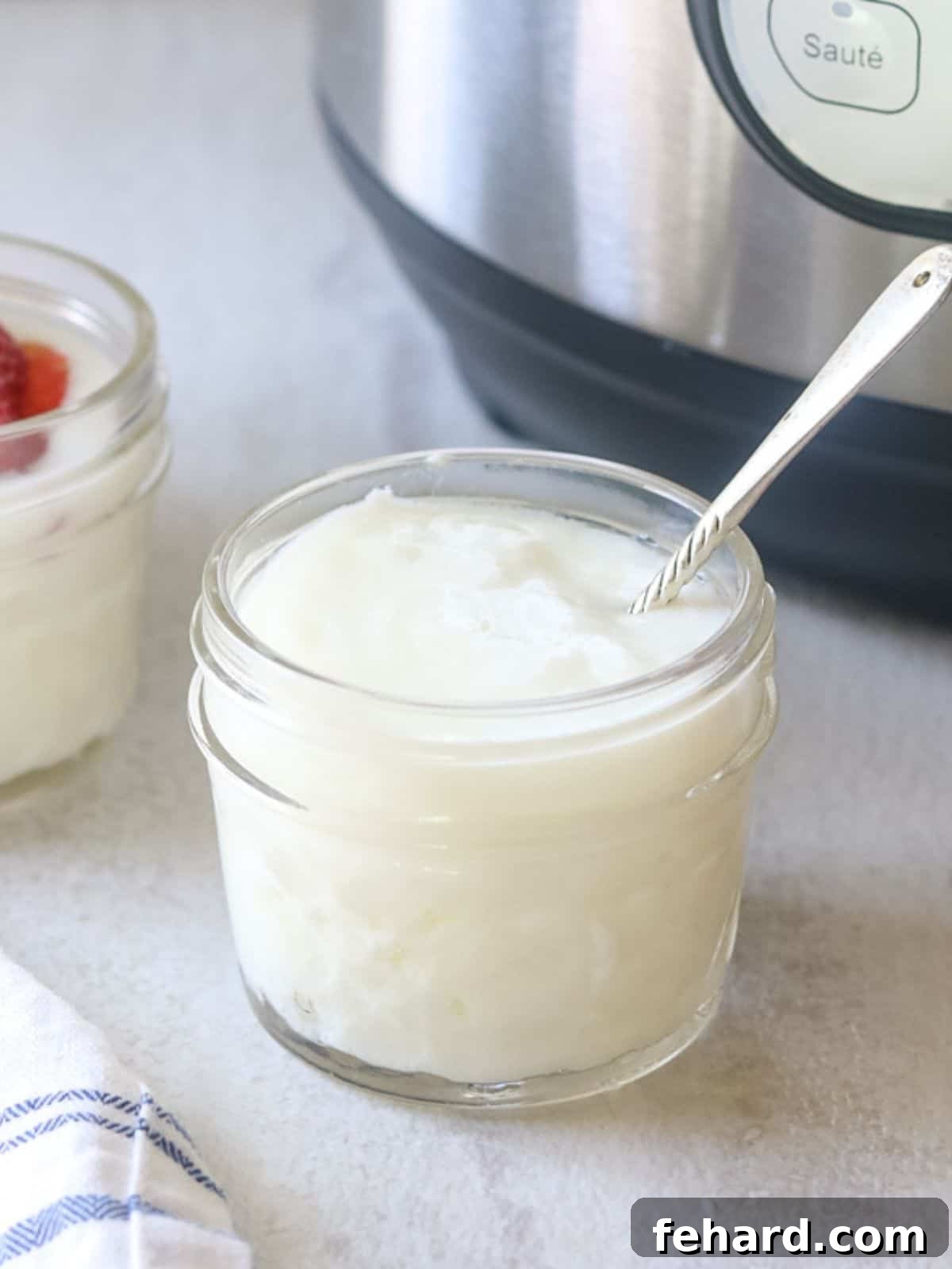 Jar of homemade Instant Pot yogurt with small spoon next to an Instant Pot. Fresh, creamy yogurt is visible.