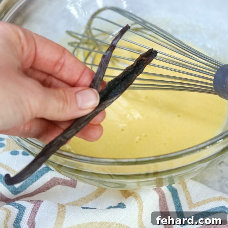 Infusing the custard with fresh vanilla bean for an authentic Creme Brulee taste. A vanilla bean pod, split open lengthwise, with its fragrant seeds being scraped into a glass mixing bowl containing the egg yolk and sugar custard mixture.