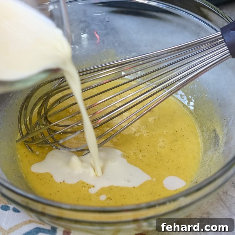 The tempering process: slowly combining warm cream with egg yolks for a perfect Creme Brulee. Warm cream being carefully and slowly poured into the vigorously whisked egg and sugar mixture in a glass bowl. A whisk is visible, ensuring continuous stirring.