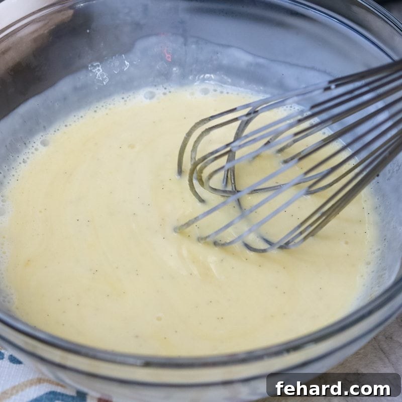 Final whisking of the creme brulee custard to ensure a perfectly smooth consistency. A metal whisk blending the smooth, pale yellow custard mixture in a clear glass bowl, indicating the custard is thoroughly combined and ready for the ramekins.