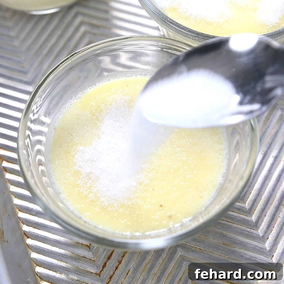 Applying the sugar topping for the signature caramelized crust. A hand sprinkling an even layer of turbinado sugar over the top of a set Creme Brulée in a ramekin, preparing it for the caramelizing step.