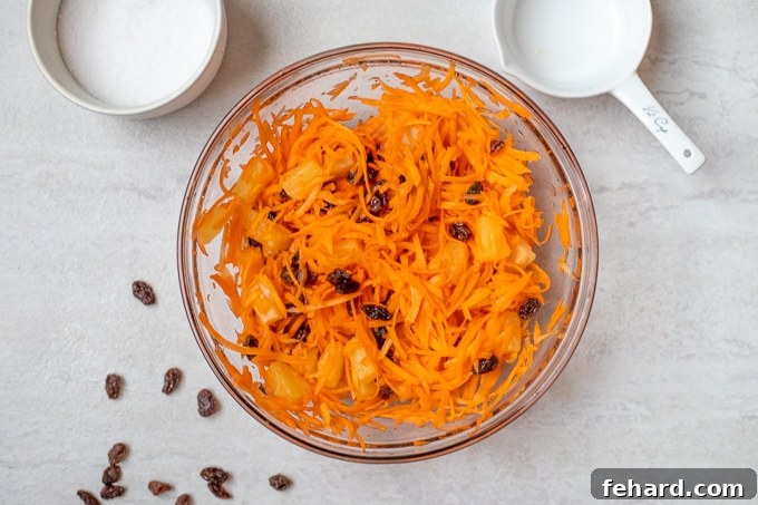 Shredded carrots, pineapple tidbits, and plumped raisins being tossed with the sweet and tangy dressing in a large mixing bowl.