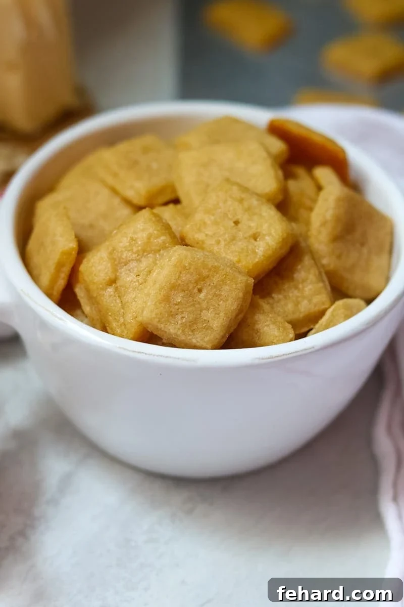 A bowl overflowing with golden brown homemade cheese crackers, with a cheese grater and fresh cheese in the soft-focus background. The crackers are perfectly square and have a distinct texture.