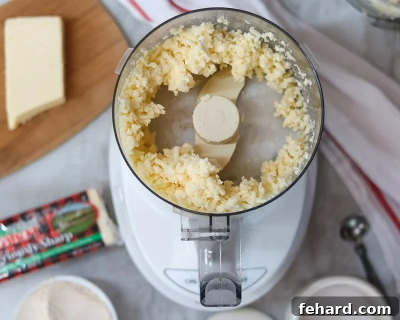 Close-up shot of freshly shredded sharp cheddar cheese and cold, cubed butter inside a food processor bowl, ready to be processed into cracker dough.