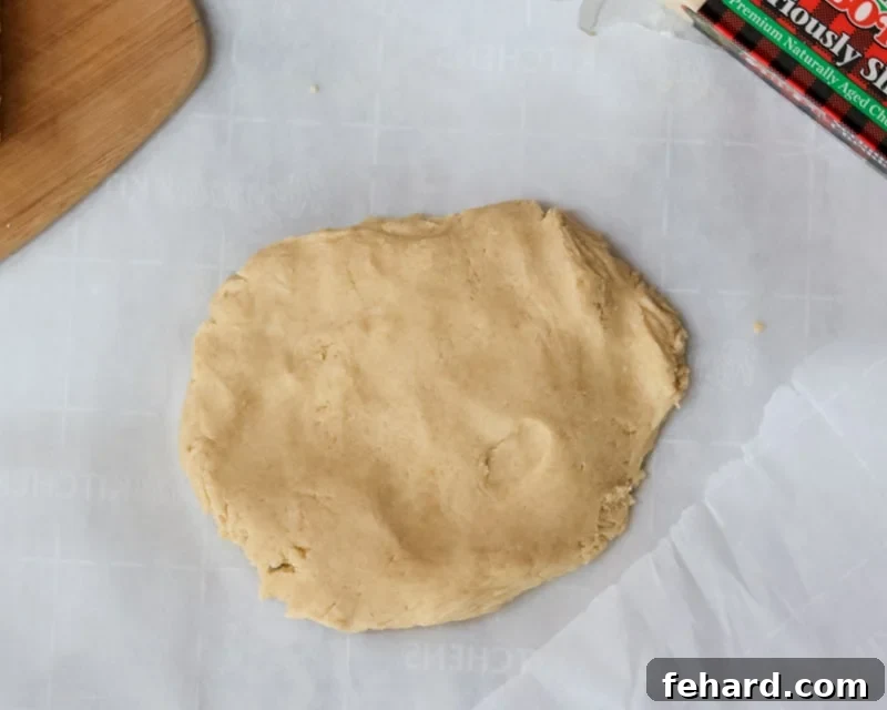 A smooth, round disk of yellow cheese cracker dough resting on a sheet of parchment paper, indicating it's ready for chilling.