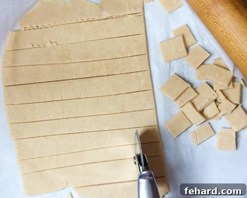 A pizza cutter being used to precisely slice rolled-out cheese cracker dough into small, even squares on parchment paper.