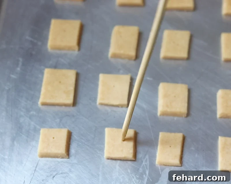 A hand using a toothpick to poke a small hole in the center of an uncooked cheese cracker square, preparing it for baking.