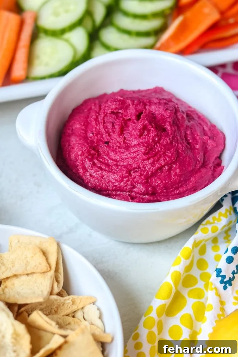 A bowl of vibrant pink roasted beet hummus garnished with fresh herbs and a drizzle of olive oil, surrounded by an assortment of colorful vegetables like cucumber slices, carrot sticks, and bell pepper strips, alongside crispy pita chips on a rustic wooden board.