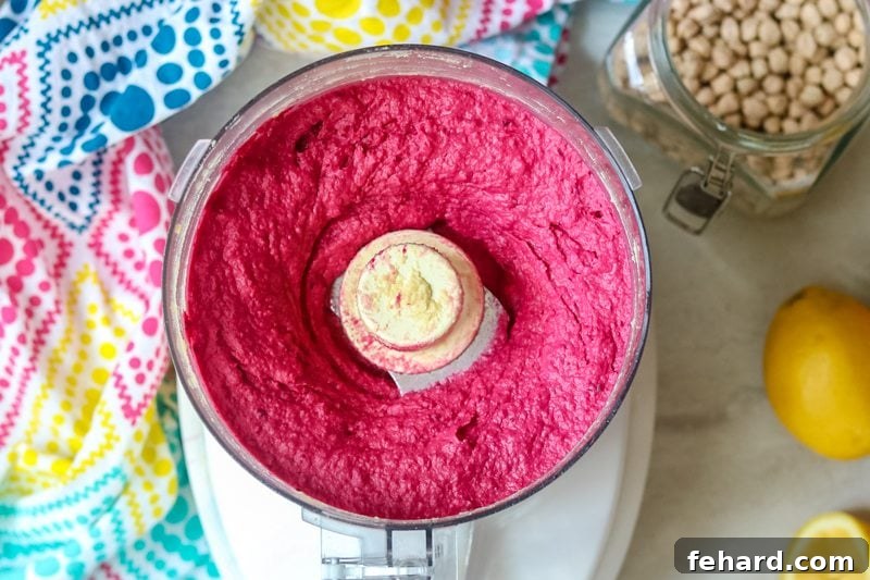 Bright pink beet hummus being processed in a food processor, illustrating its smooth texture and vibrant color. The s-blade is visible, actively blending the ingredients.