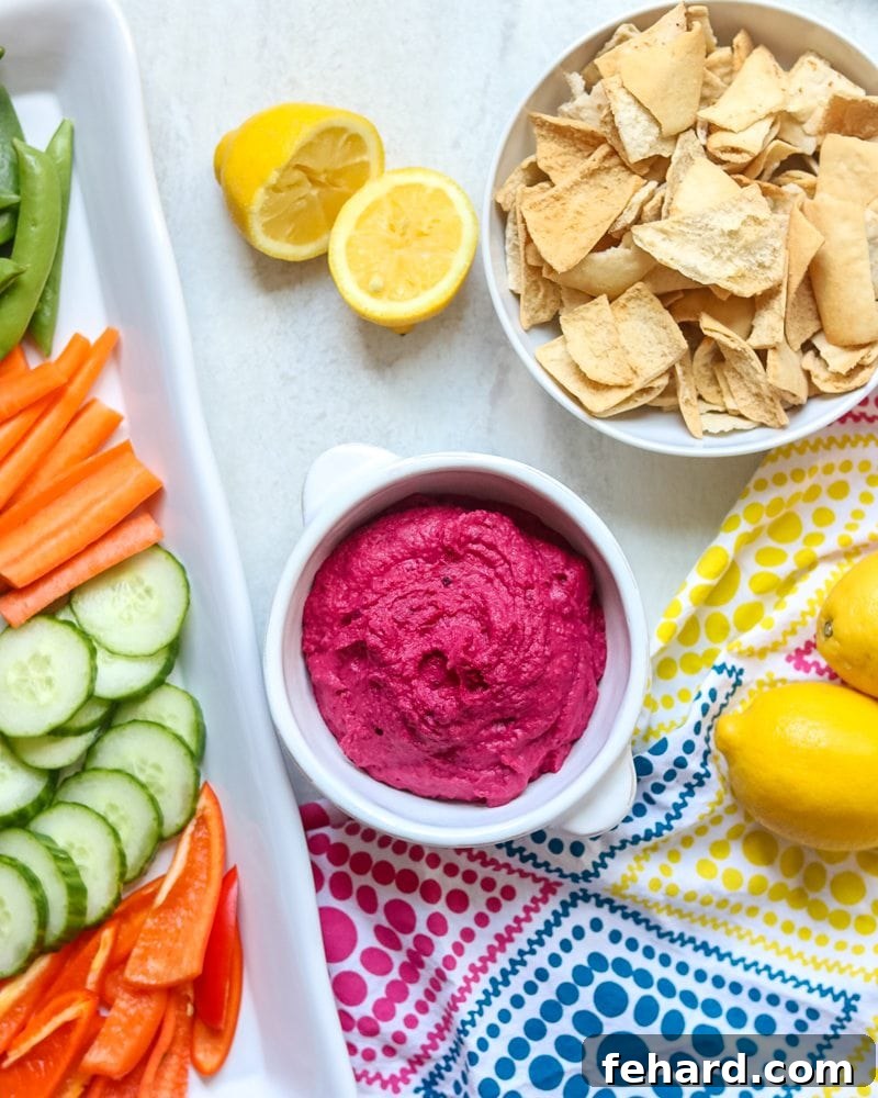 A beautifully presented serving of roasted beet hummus in a white bowl, surrounded by an appetizing selection of fresh vegetables including cucumber slices, carrot sticks, radishes, and colorful bell pepper strips, along with crispy pita chips, all arranged on a light background.