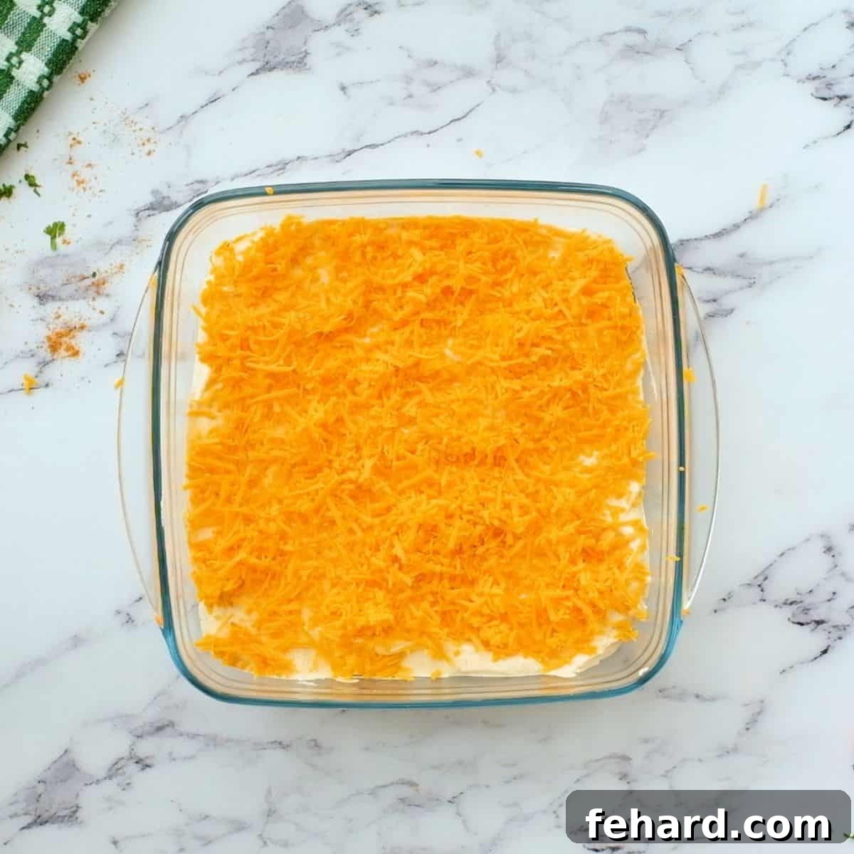 Layered taco dip with shredded cheese on top in a glass baking dish, before baking.