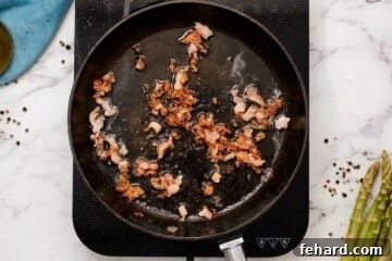 Bacon cooking in a skillet next to fresh asparagus.