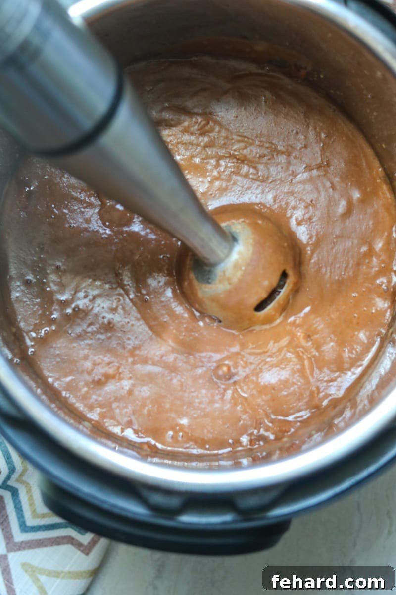 An immersion blender actively blending cooked pinto beans directly in the Instant Pot, creating creamy refried beans.