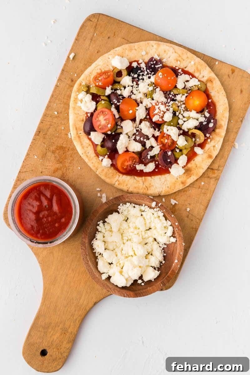 An assembled Pita Pizza generously topped with crumbled feta cheese, black olives, and sliced tomatoes, ready for baking.