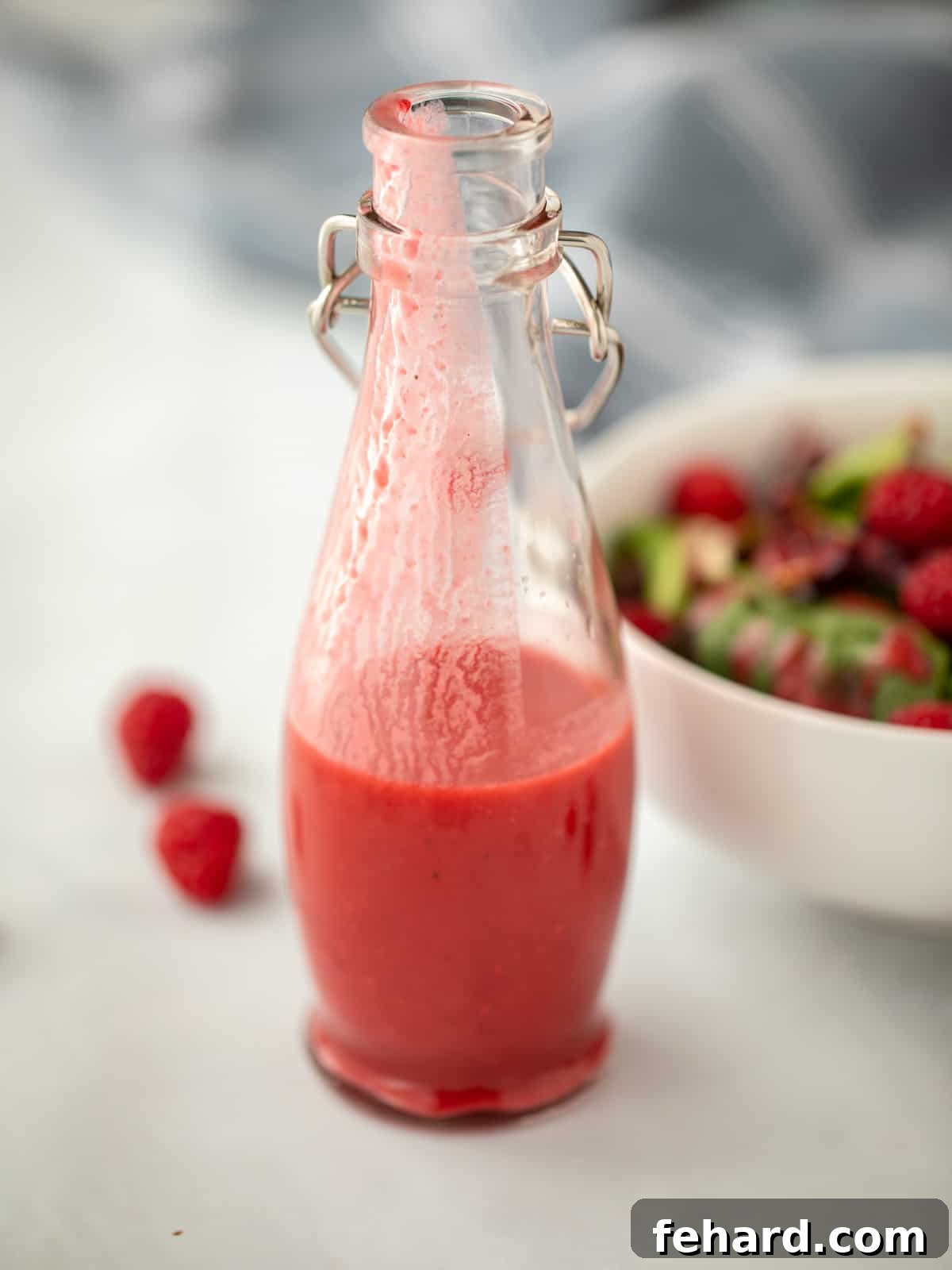 Bottle of homemade raspberry vinaigrette dressing next to a fresh green salad topped with raspberries.