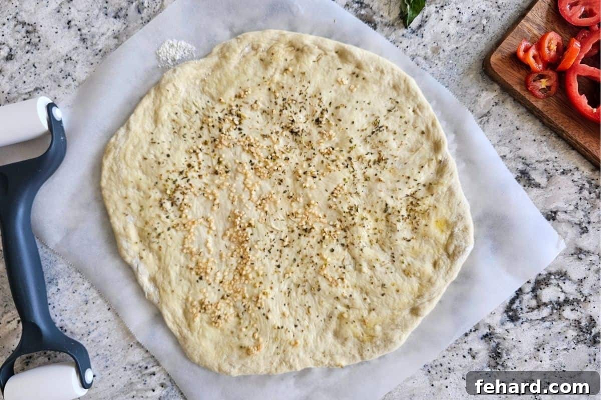 Pizza dough rolled out on parchment paper, brushed with olive oil, and seasoned with minced garlic and oregano, ready for toppings.