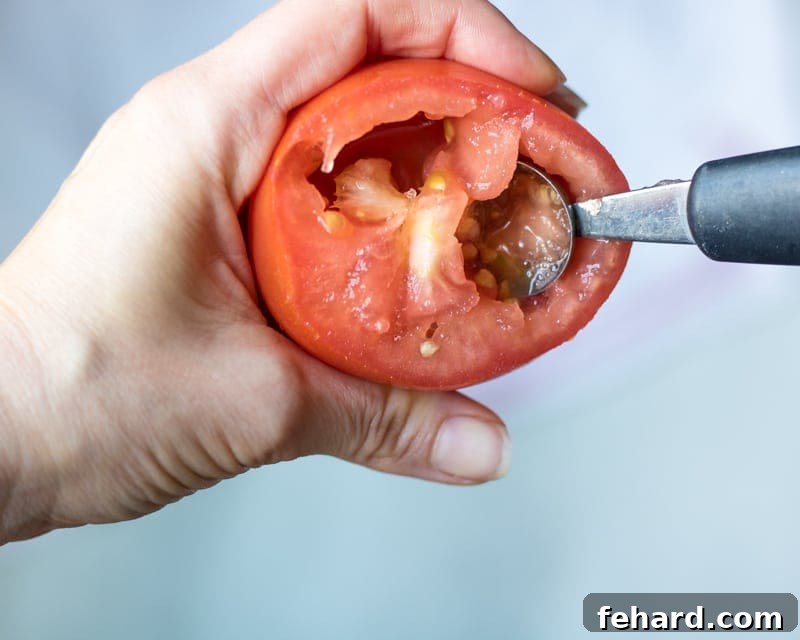 A hand using a melon baller to gently remove seeds from a halved tomato, a crucial step for a crisp pizza.