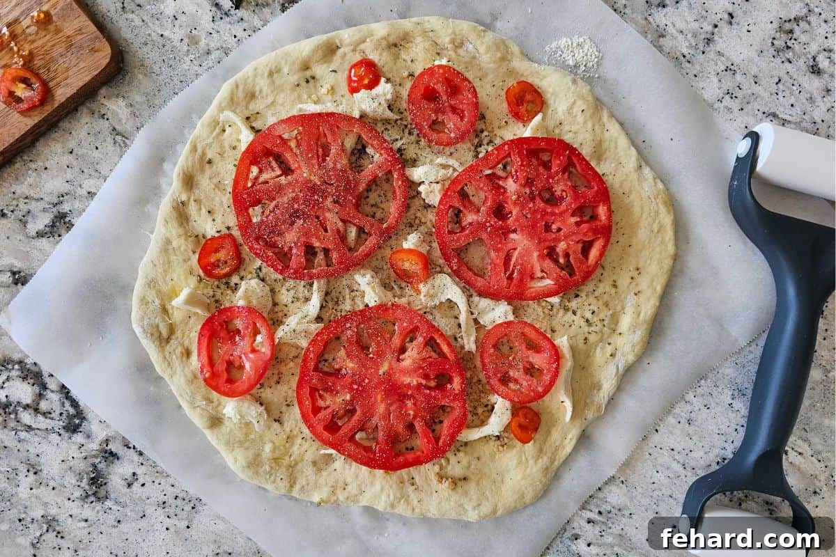 Prepared pizza dough with olive oil, seasonings, fresh mozzarella, and Parmesan cheese, now topped with seasoned fresh tomato slices before baking.