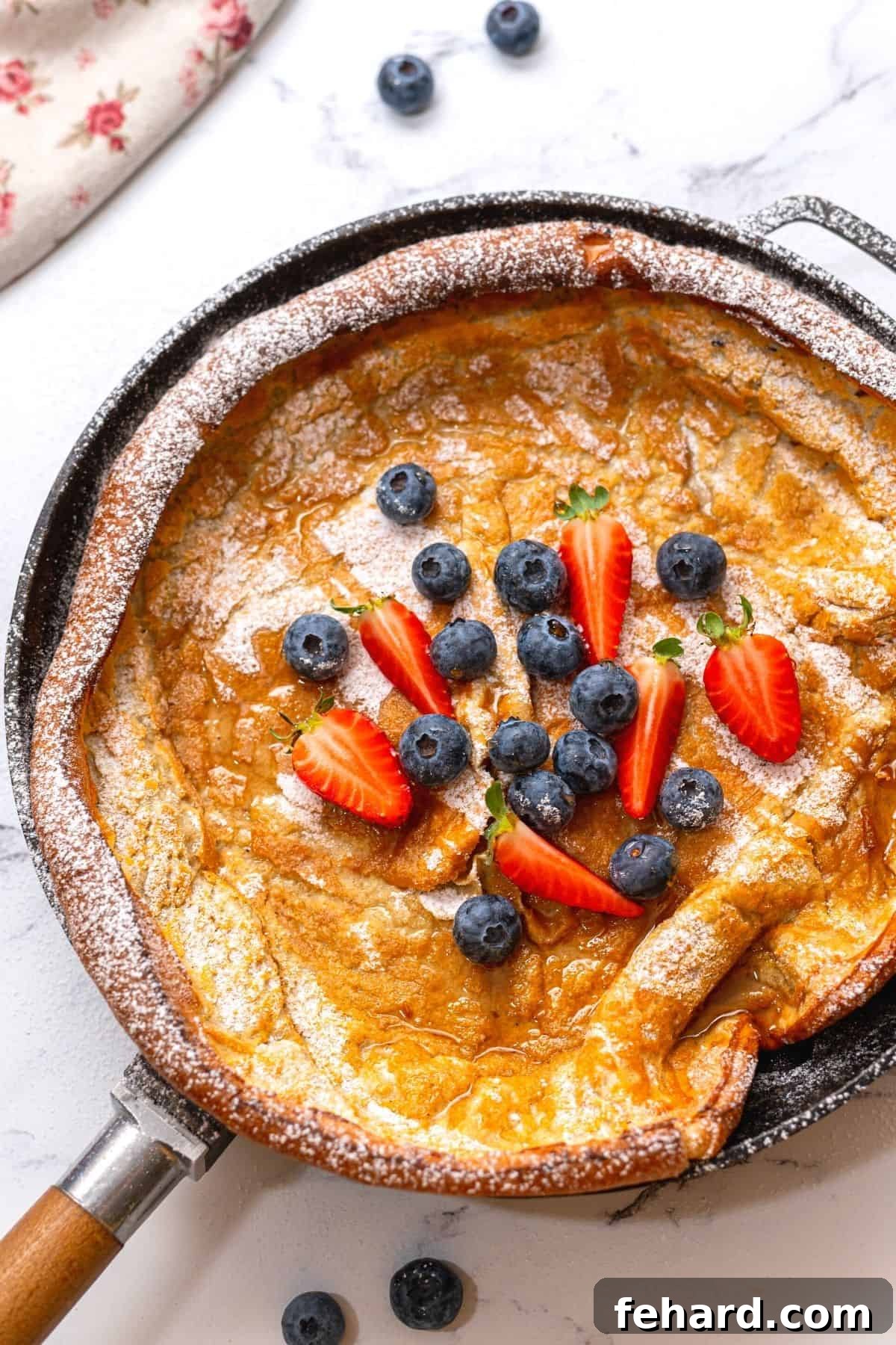 Golden brown Dutch Baby Pancake in a cast iron skillet, generously topped with fresh berries and a dusting of powdered sugar, fresh from the oven.