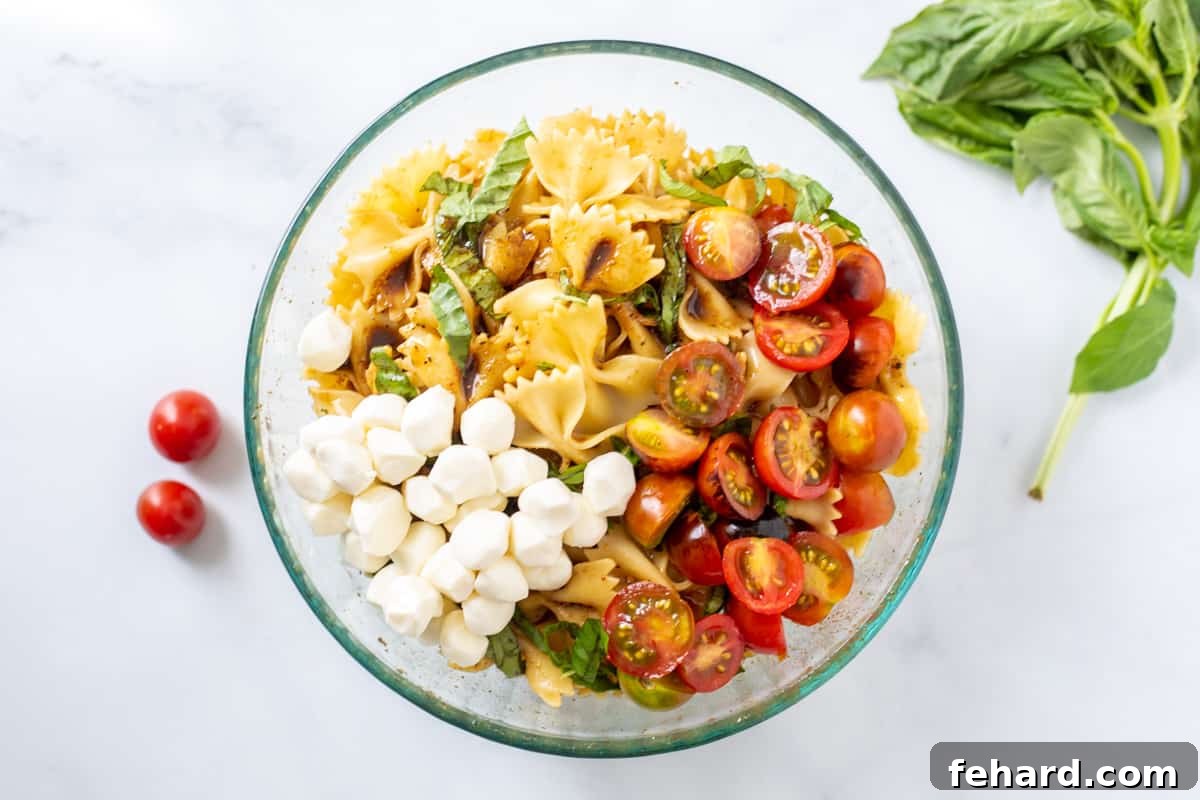 A large mixing bowl containing tossed pasta, fresh mozzarella pearls, halved grape tomatoes, and basil ribbons, showcasing the final stages of Caprese Pasta Salad preparation.