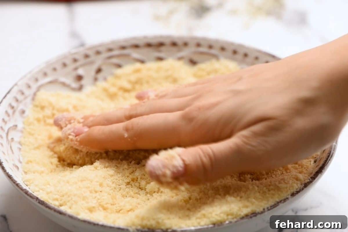 Oven Baked Chicken Tenders Breading A hand gently pushing chicken strips into a bowl of Panko breading to ensure an even and crispy coat.