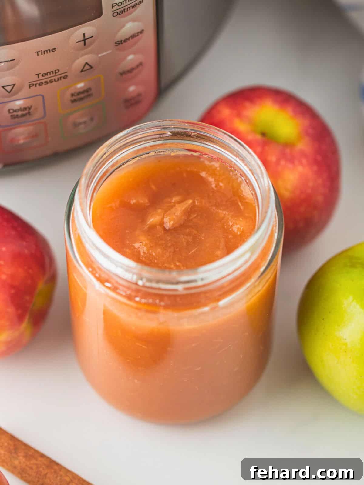 Jar of homemade Instant Pot applesauce next to fresh apples and cinnamon sticks, with an Instant Pot in the background.