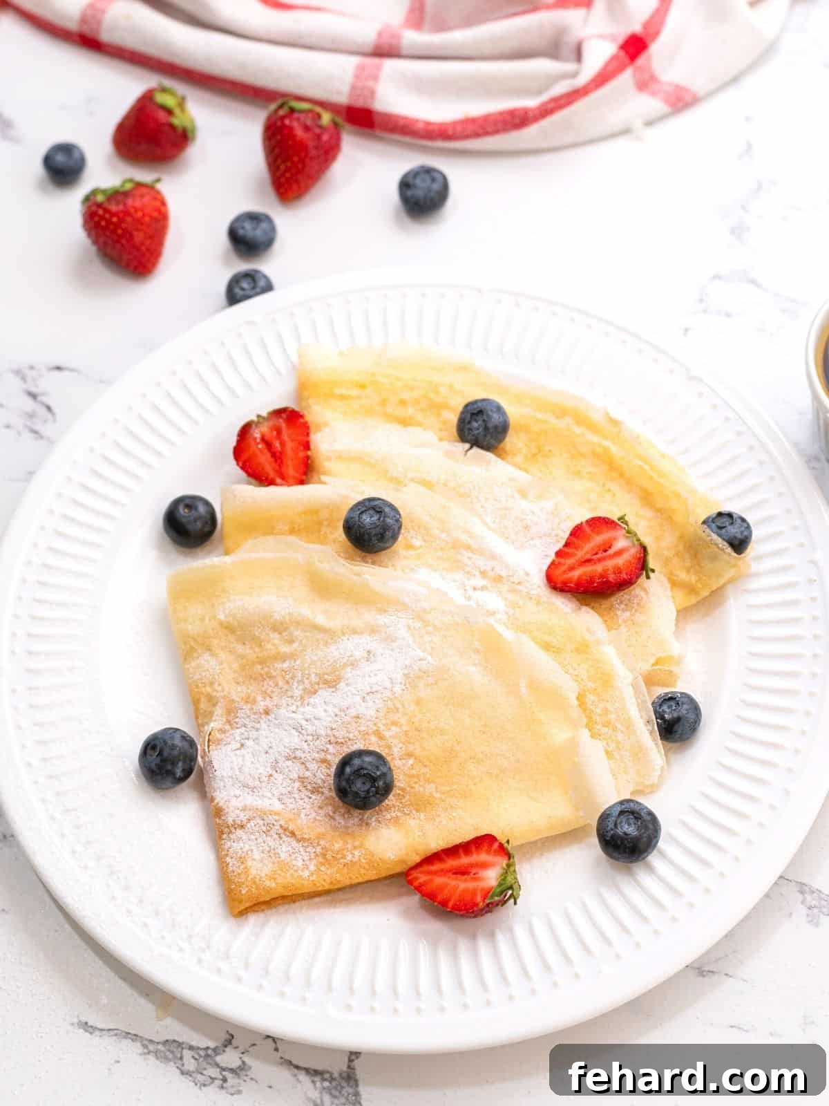 Four golden-brown crepes delicately folded on a white plate, beautifully topped with a dusting of powdered sugar and a vibrant medley of fresh berries.