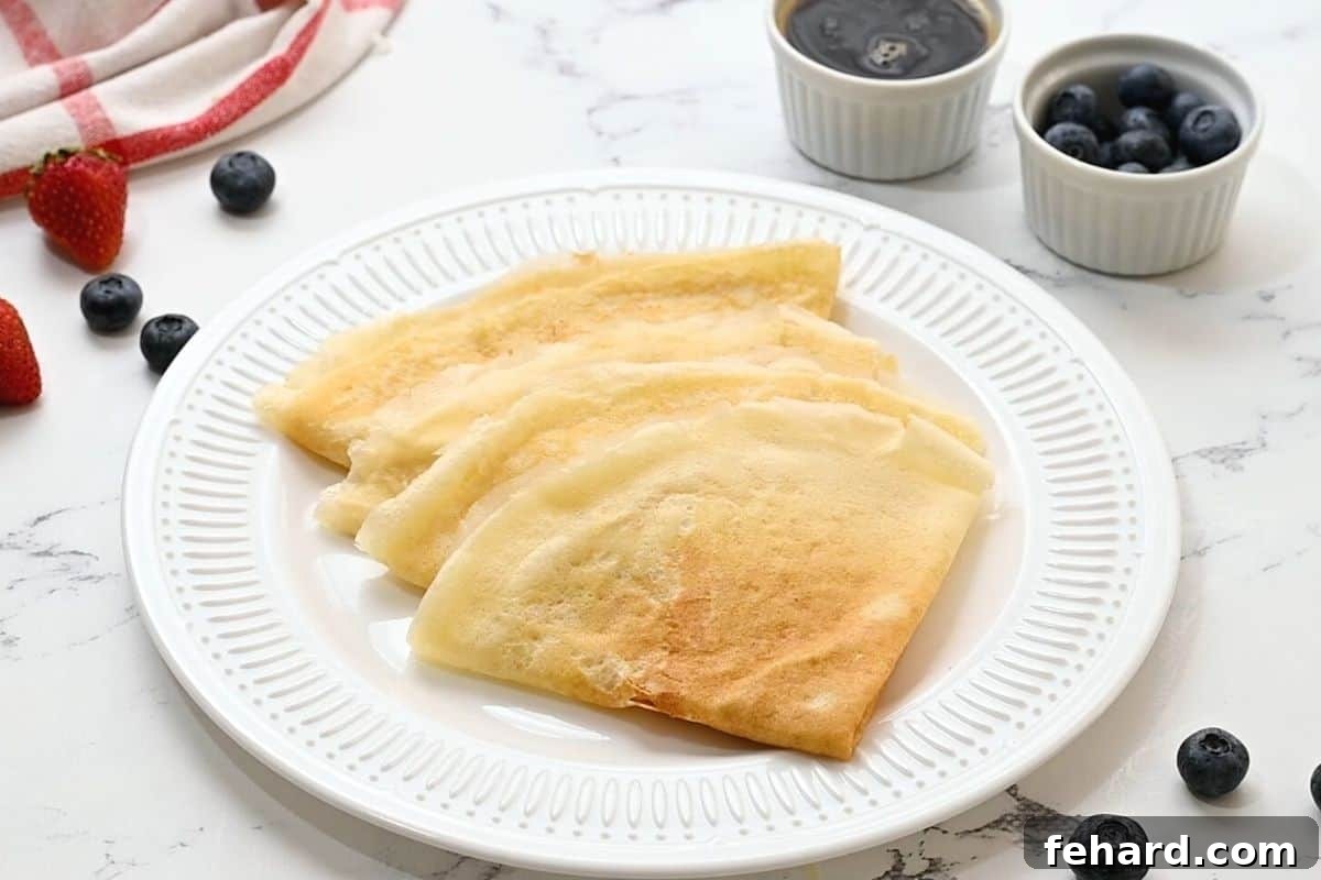 Four perfectly folded golden crepes arranged on a white plate, accompanied by a small pitcher of rich maple syrup, ready for serving.