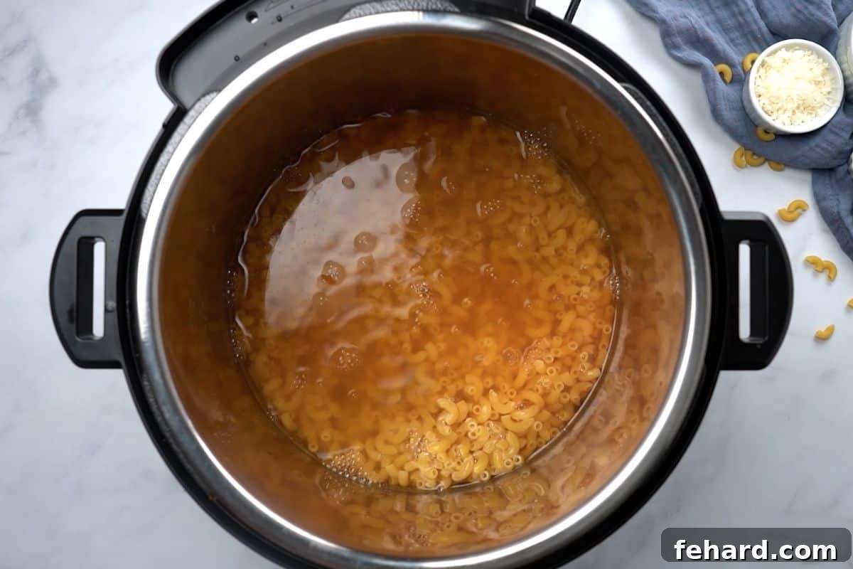 Elbow noodles are perfectly submerged in a richly seasoned broth inside the Instant Pot's inner pot, ready for pressure cooking to create delicious mac and cheese.