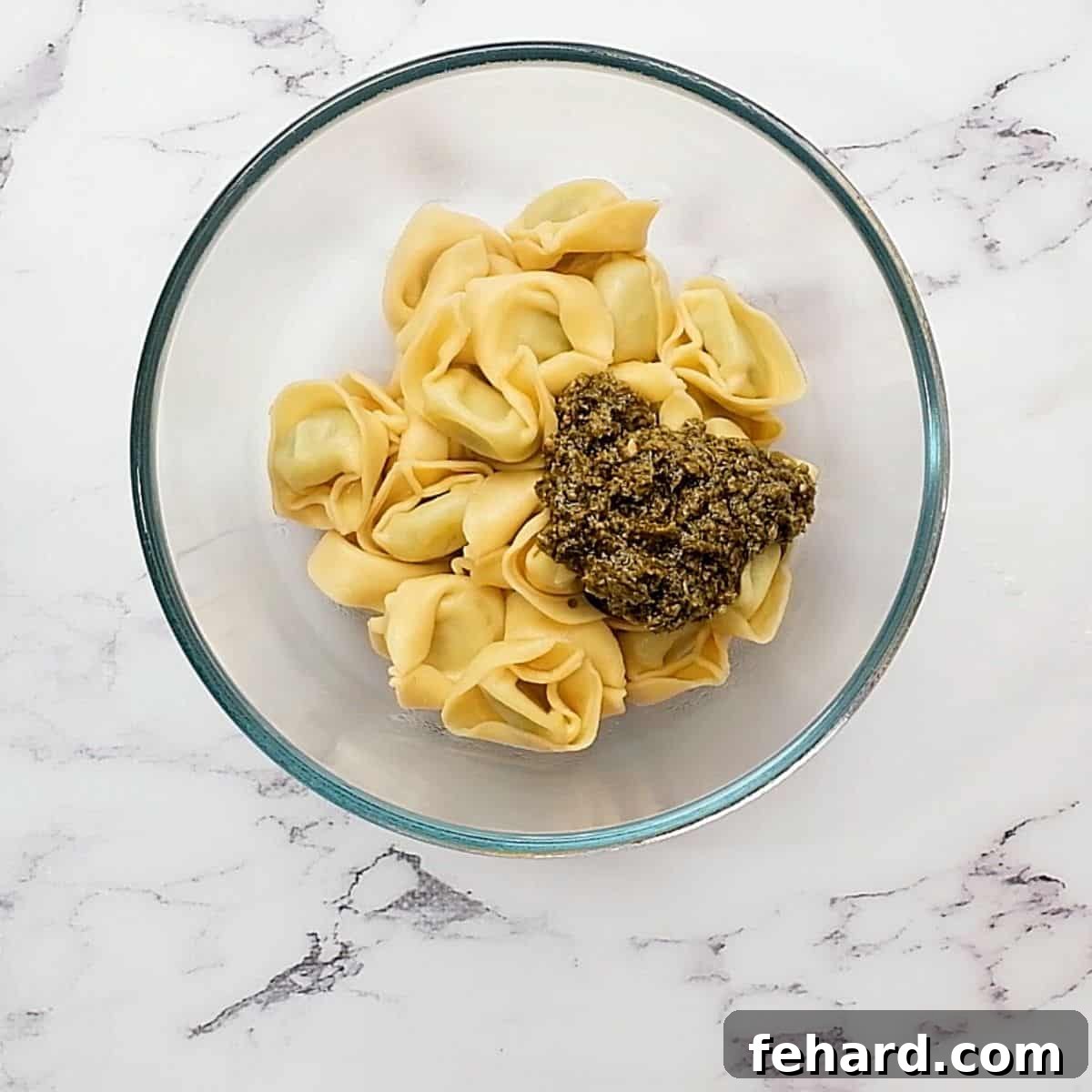 Warm cheese tortellini mixed with vibrant green pesto in a glass mixing bowl, ready for cooling.