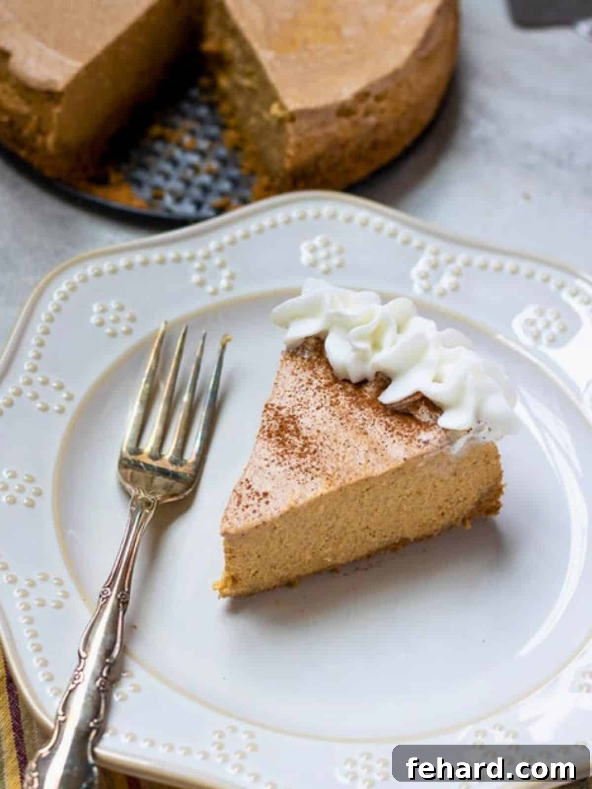 A perfectly sliced piece of Instant Pot Pumpkin Cheesecake on a white plate, garnished with homemade whipped cream and a fork. The rest of the whole cheesecake is blurred in the background, showcasing its smooth, creamy texture and golden-brown gingersnap crust.
