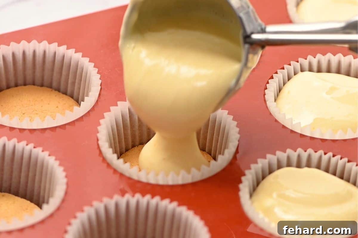 A small cookie scoop gently drops creamy cheesecake batter into a mini muffin pan lined with vanilla wafer crusts, ensuring even portioning.