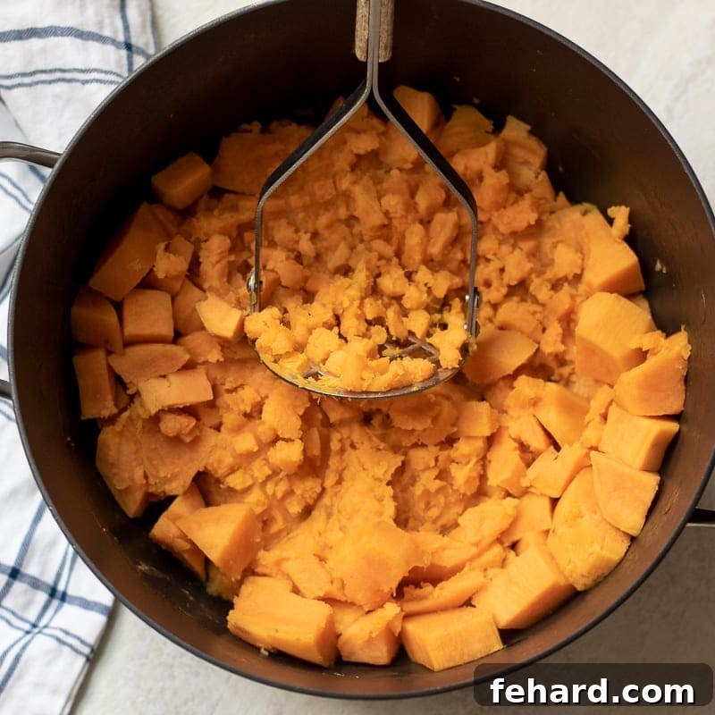 Healthy Mashed Sweet Potato Recipe A potato masher actively mashing cooked sweet potatoes in a large pan, creating a smooth and creamy texture.