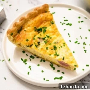 Slice of ham and cheese quiche topped with chopped chives on white plate.