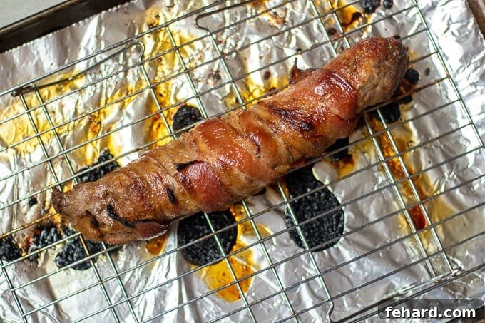 Oven Roasted Pork Tenderloin wrapped in bacon on Baking Sheet, with a thermometer inserted.