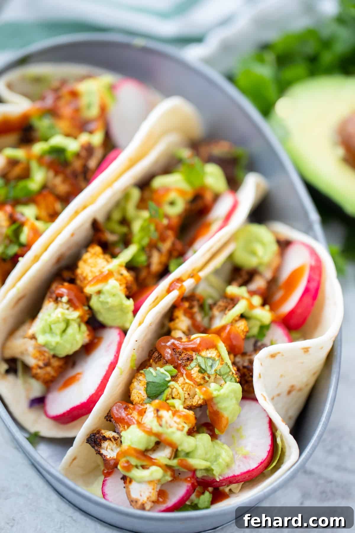 Cauliflower Tacos Three tortillas topped with golden roasted cauliflower and a generous drizzle of avocado cream, elegantly arranged next to fresh avocado halves, highlighting the vibrant colors and textures of the dish.
